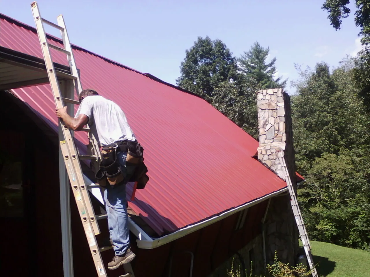 Roofer on a ladder beside a red metal roof and stone chimney — Soffit & Fascia Repair by Roofing Brothers in Edinboro